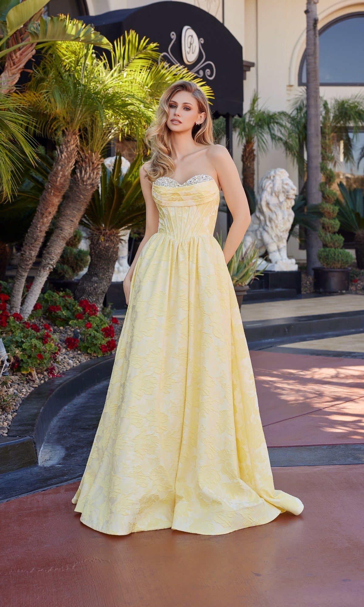 A woman wearing the yellow Nox Anabel T1912 long prom dress with a jeweled sweetheart neckline stands outdoors by palm trees and plants, a building entrance in the background.